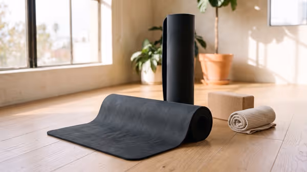 A plain charcoal yoga mat partly unrolled on a light oak floor in a sunlit studio, with a cork block and a rolled towel beside it — an illustration of the mat category, not a photograph of the Alo Warrior