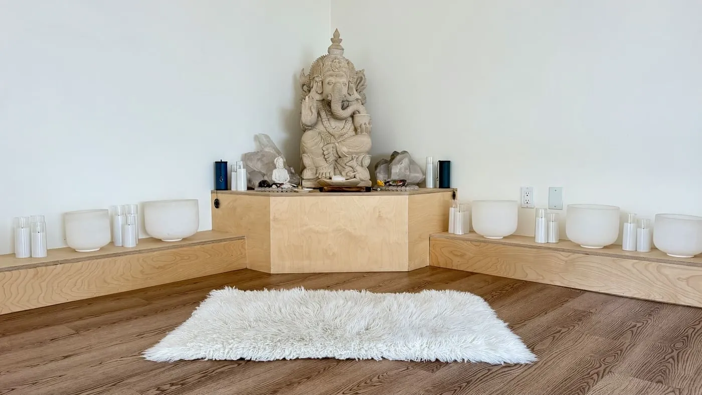 The altar corner: a carved stone Ganesha on a pale wooden plinth, rows of white crystal singing bowls and candles on low steps either side and a sheepskin rug laid on the floor in front
