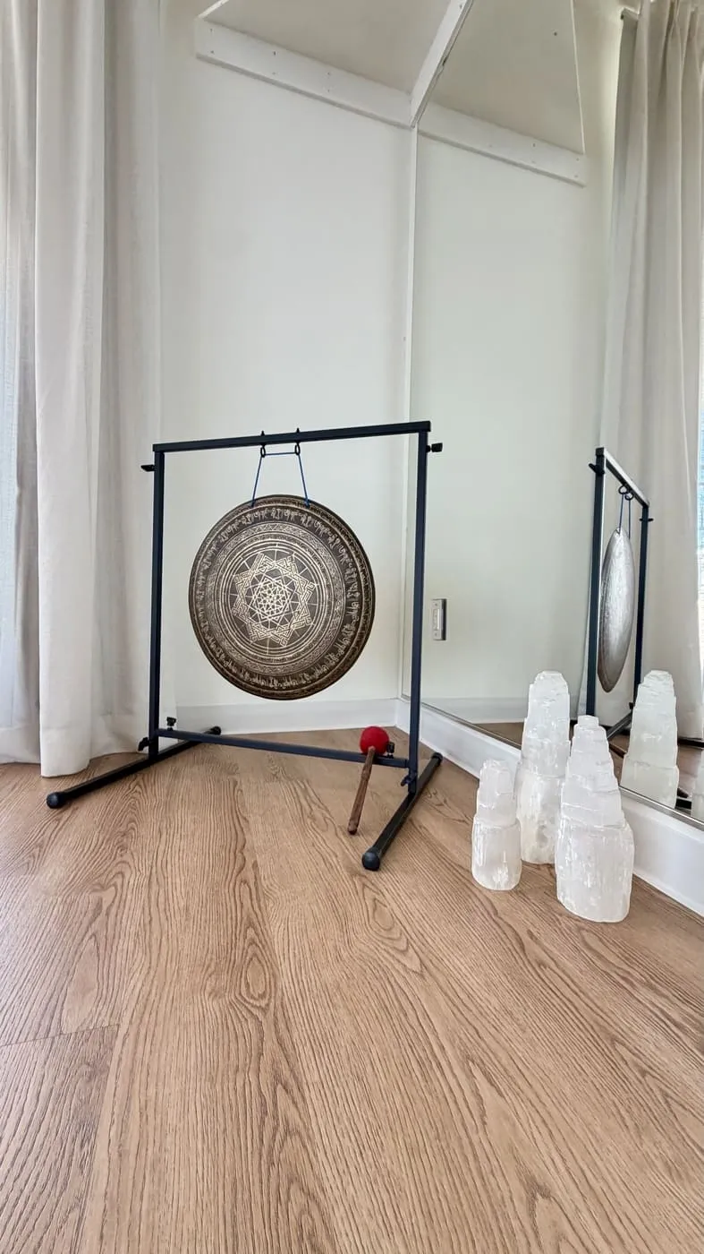 A hanging gong on a black stand beside sheer curtains, with a mallet and a few white salt lamps on the wood floor