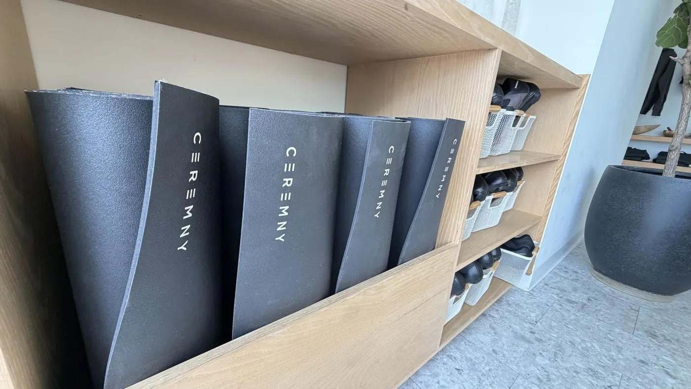 Black rolled yoga mats printed with the CEREMNY name, standing in a pale wooden cubby with grey bins of accessories beside them