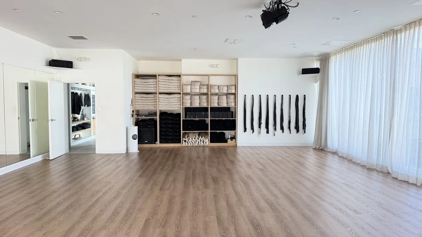 The practice room facing the prop wall: a pale wooden cubby unit of folded blankets and black bolsters, a row of black straps on hooks, a mirror and sheer curtains down the right-hand side