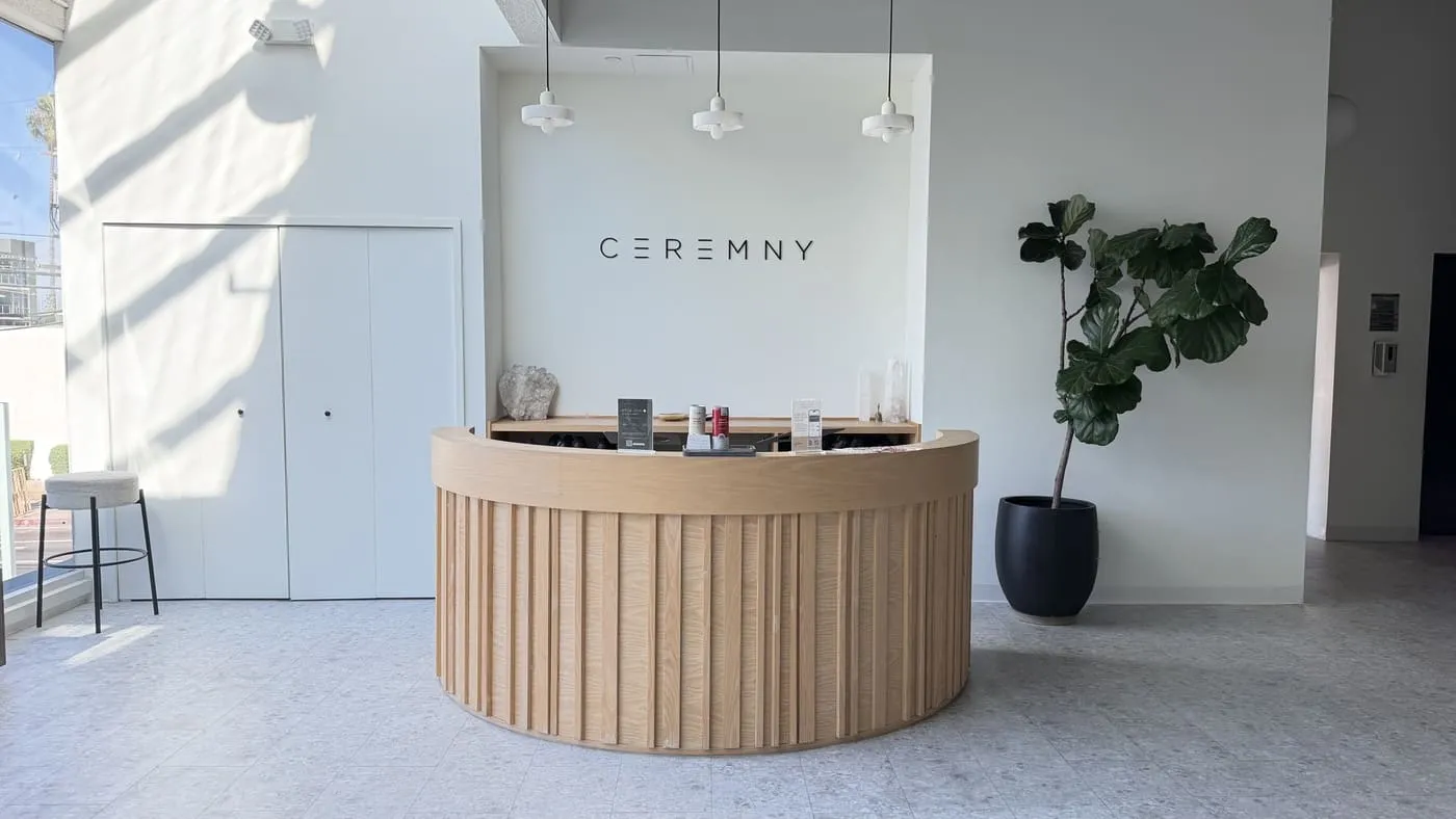 The front of house: a curved pale-wood reception desk under a cluster of white pendant lights with the CEREMNY name on the wall behind, a fiddle-leaf fig beside it and a bar stool at the window