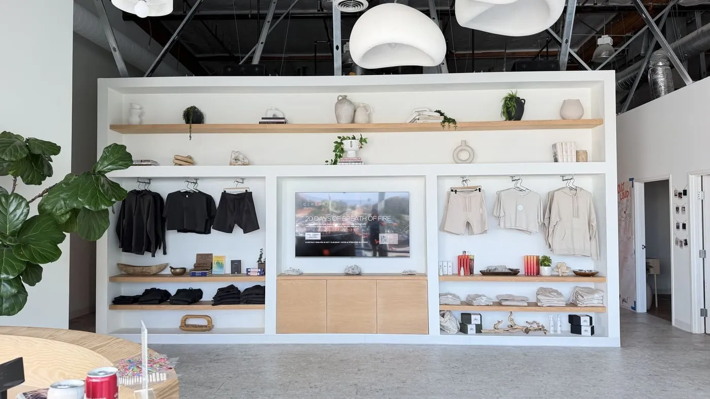 The retail wall beside reception: built-in white shelving with folded and hung clothing, ceramics and a wall-mounted screen under white pendant lights