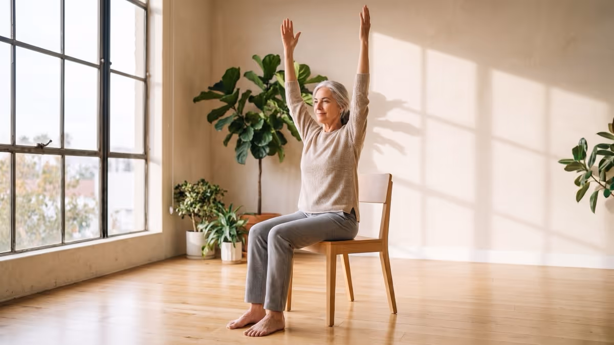 A silver-haired woman in her seventies sitting tall on a plain wooden dining chair with no wheels or armrests, both feet flat on the floor, reaching both arms straight overhead in a sunlit studio