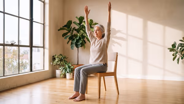 A silver-haired woman in her seventies sitting tall on a plain wooden dining chair with no wheels or armrests, both feet flat on the floor, reaching both arms straight overhead in a sunlit studio