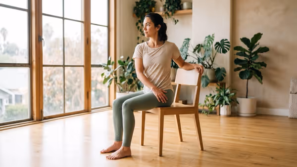A woman in her forties practising a seated twist on a plain wooden dining chair in a sunlit studio, both feet flat on the floor, one hand on the chair back behind her and the other on her thigh — no mat, and a chair with no wheels or armrests