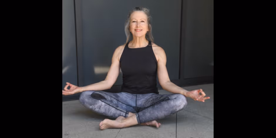 Photograph of yoga teacher Kate O'Neal sitting cross-legged on a concrete floor in a black tank top and grey leggings, hands resting on her knees, smiling, in front of a dark wall