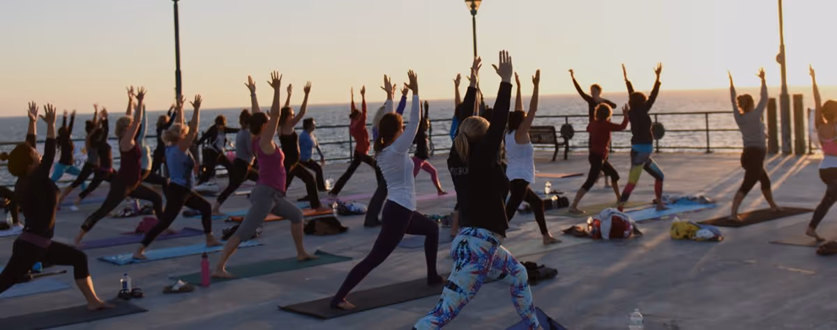 A large outdoor yoga class on the pier deck at sunset, seen from behind, everyone in a high lunge with arms raised over mats, with lamp posts, the railing and the ocean beyond