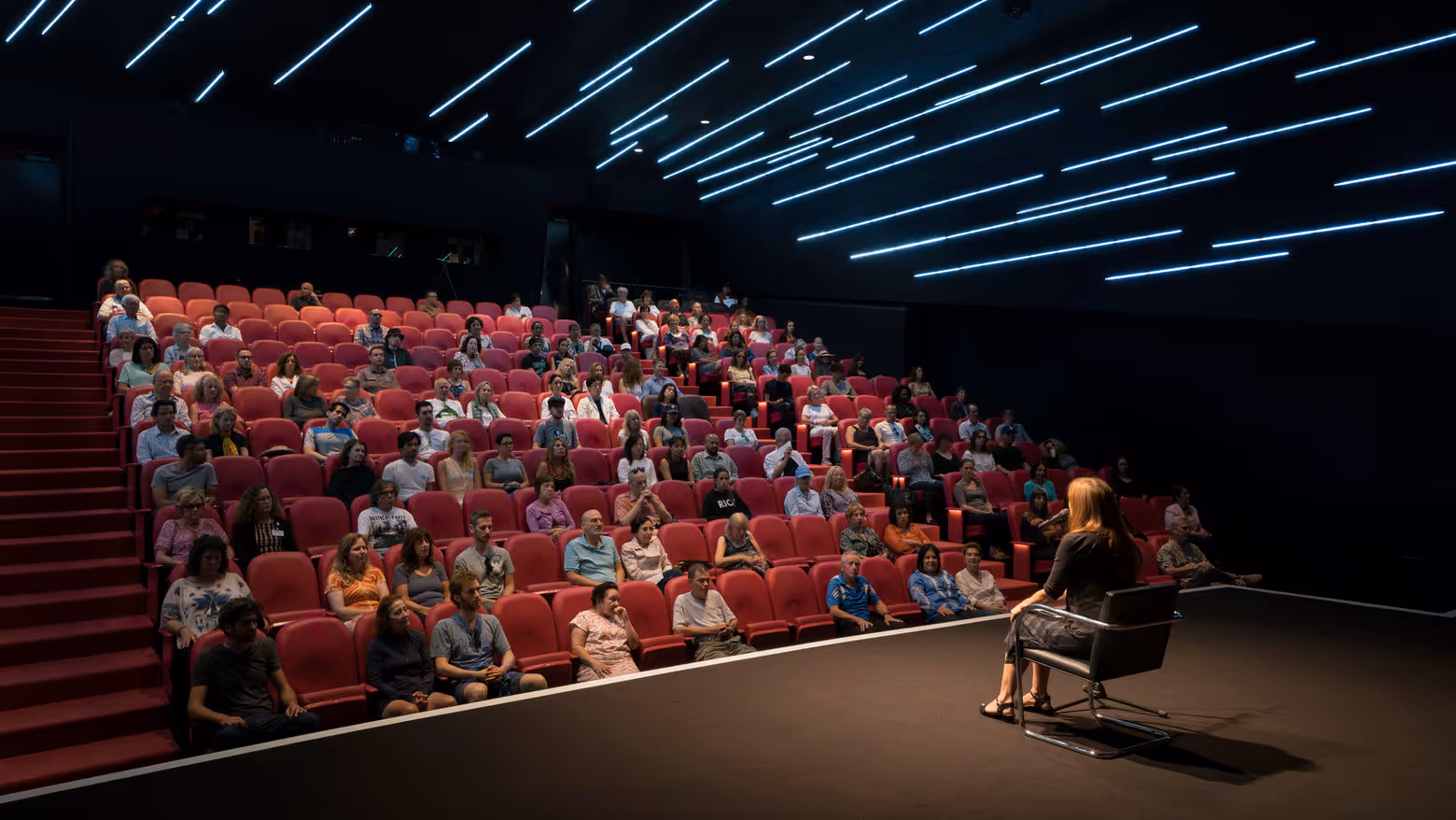 Photograph from the Hammer Museum: a teacher seated on a chair at the edge of the Billy Wilder Theater stage, facing an audience in rows of red seats under streaks of ceiling light