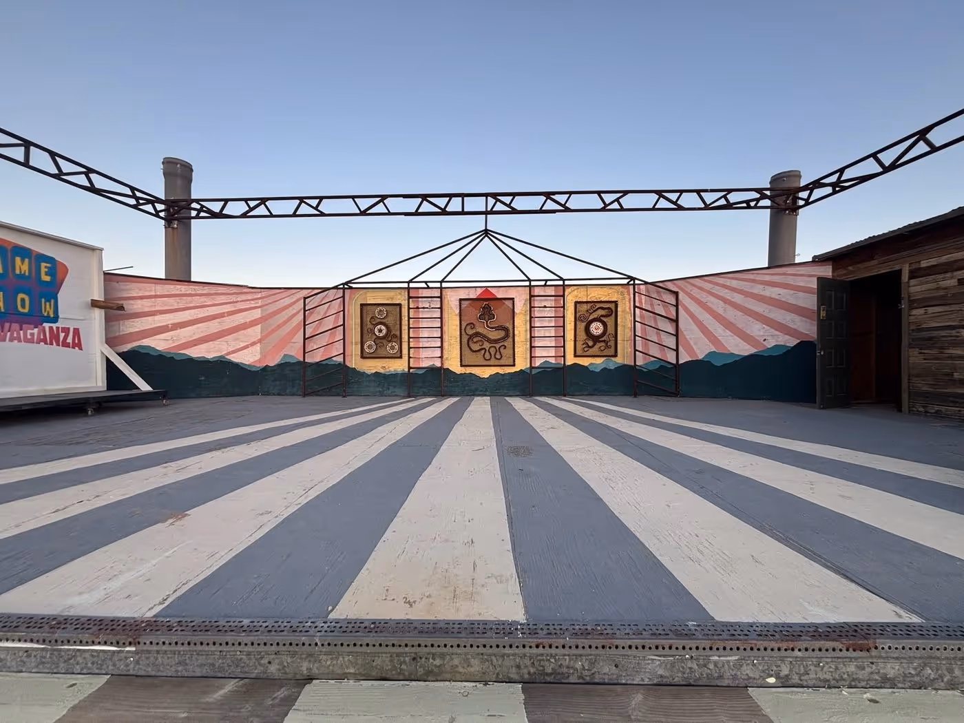 Photograph of the festival grounds: an empty outdoor stage with a blue-grey and cream striped floor, a pink sunburst backdrop painted with desert mountains and three framed relief panels, and a steel truss against a clear blue sky