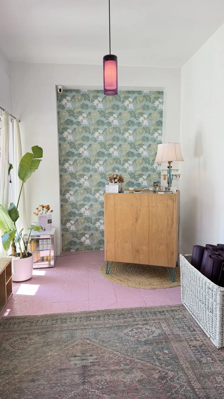 The entrance, a pink floor with a vintage rug, a wooden reception cabinet with a lamp in front of a wall of lotus-pattern wallpaper, a pendant light and a basket of rolled purple mats
