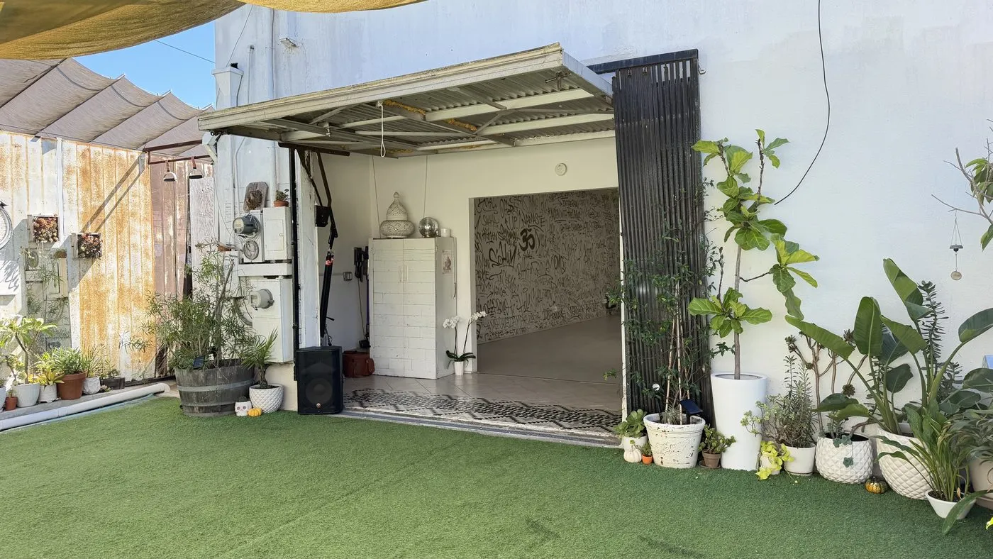 The back garden, artificial turf and potted plants under shade sails, looking into the studio through a wide open roll-up door
