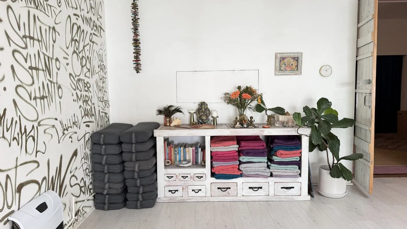 A corner of the main room, stacked grey bolsters beside a low white cabinet of folded blankets and books, with flowers, a small statue and a fiddle-leaf fig, next to the hand-lettered wall