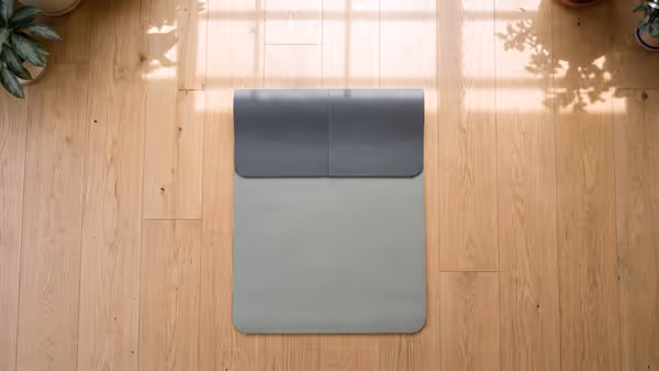 Seen from directly above, a reversible yoga mat on a light oak floor with its top third folded back so both faces show at once: a smooth sage-grey polyurethane side for sweat and a pebbled charcoal rubber side for dry practice — an illustration of the mat this guide describes, not a photograph of it