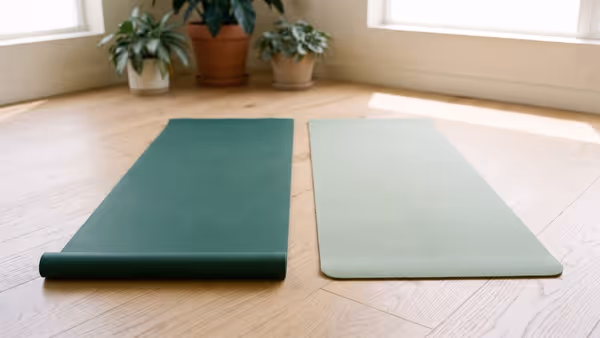 Two plain yoga mats side by side on a light oak floor, one thick and deep green, one thinner and pale sage — an illustration of the two mat types, not photographs of the named products