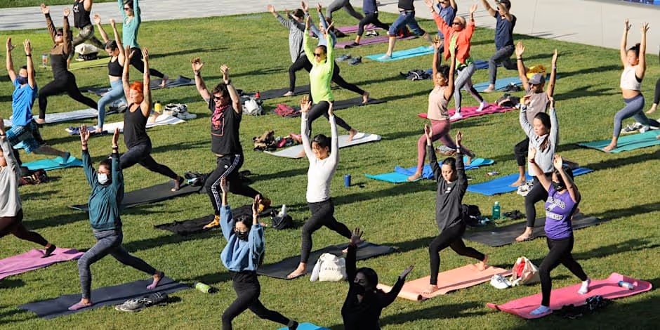 Photograph of a large outdoor yoga class on a lawn: rows of people on colourful mats in a warrior pose with arms raised, bags and water bottles beside them, in late afternoon sun