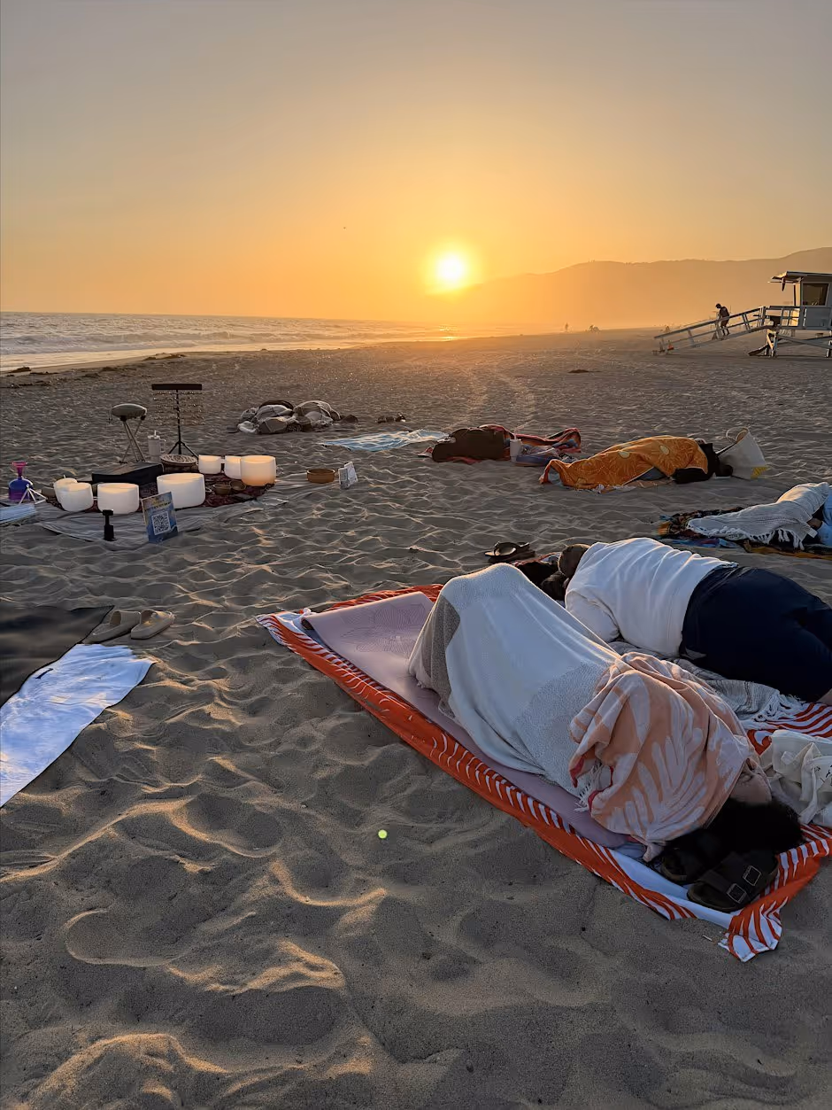 A sound bath on the sand at sunset, people lying under blankets on towels while a row of glowing white crystal singing bowls and a chime stand sit to one side, the sun low over the hills and a lifeguard tower in the distance