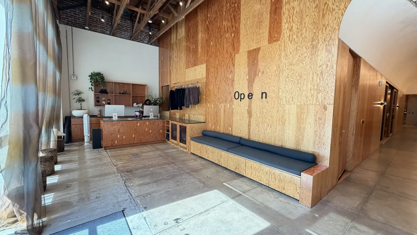 The lobby, a plywood wall lettered Open above a long upholstered bench, a rail of studio clothing and a reception desk with plants beside a tall window hung with sheer curtains