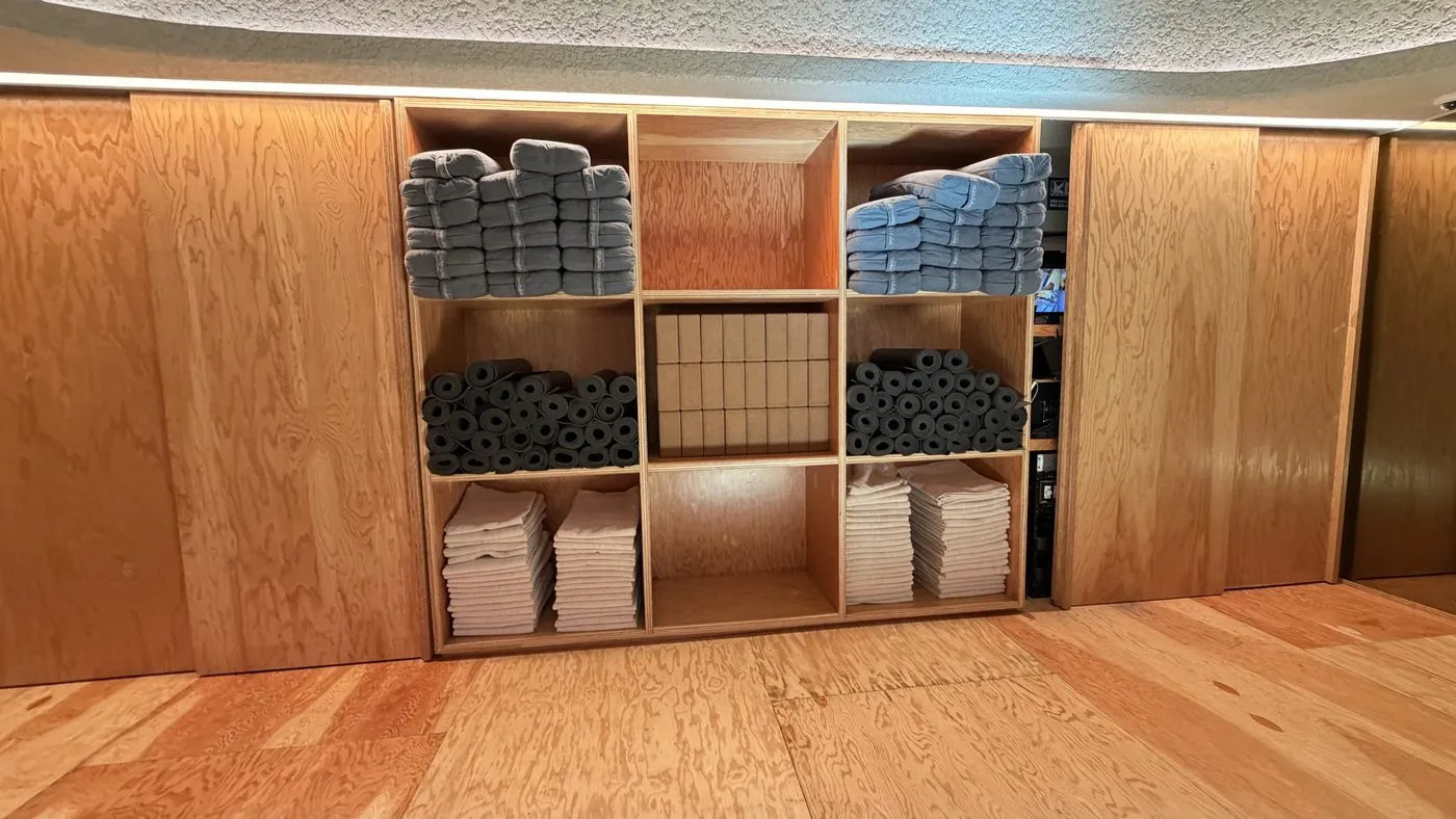 A plywood cubby unit set into the wall, holding stacks of folded grey blankets, rolled black mats, white towels and blocks