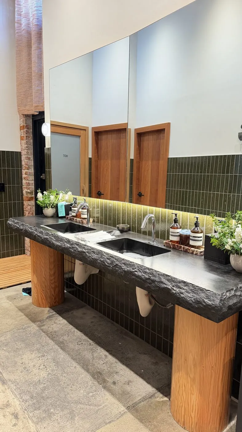 The washroom, two sinks set into a rough-edged black stone counter on round wooden legs under a long mirror and a wall of dark green tiles, with soap bottles and a vase of white flowers