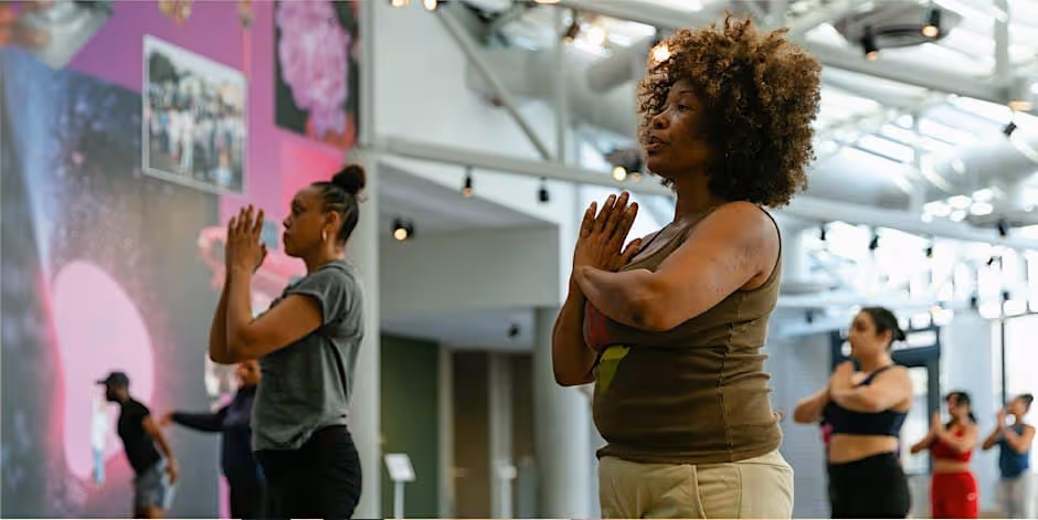 Photograph of a yoga class inside the museum: women standing with palms pressed together at the chest, eyes on the teacher, under a white industrial ceiling beside a pink and purple mural