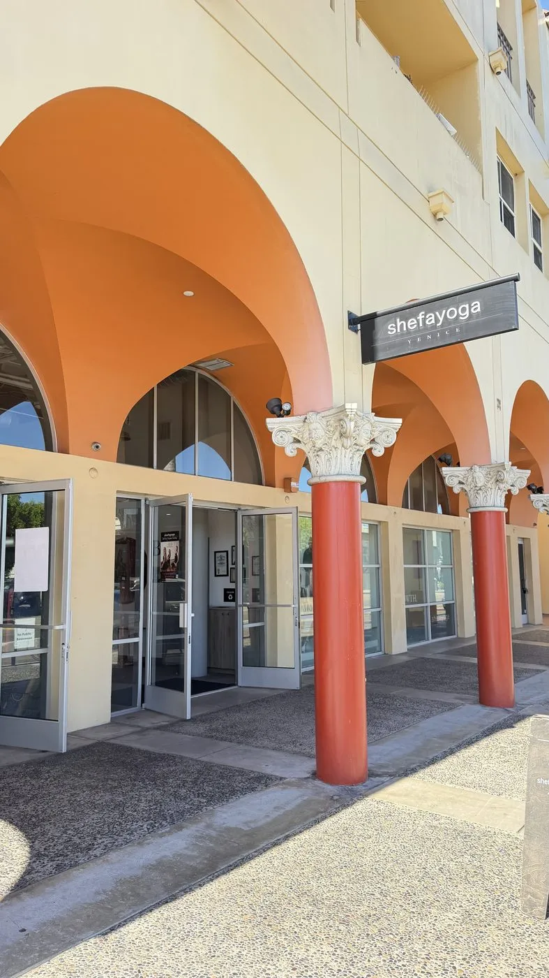 The entrance under an arcade of orange arches and red columns with carved white capitals, a hanging sign reading shefayoga Venice above the open glass doors