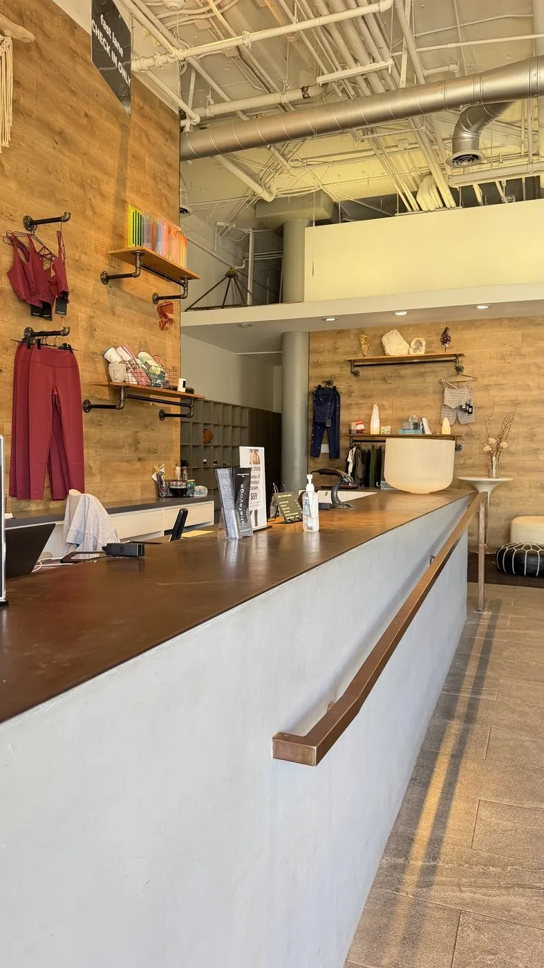 The front desk, a long concrete counter with a wooden top and a copper handrail, with studio clothing and a row of colourful yoga blocks on shelves against a reclaimed-wood wall