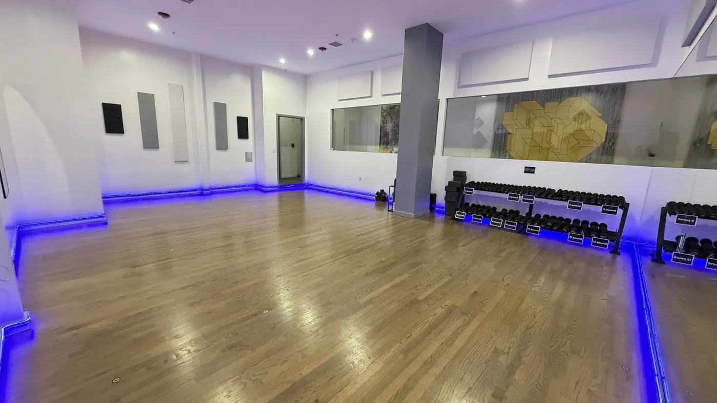 The same room from another corner, a square grey column near the back and racks of hand weights along the right wall, with the wood floor and blue floor-edge lighting running round the room