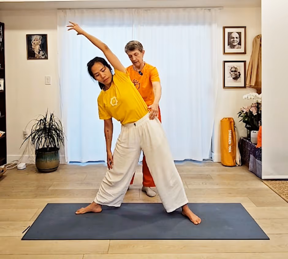 Photograph from Sivananda Yoga Los Angeles: a teacher in orange stands behind a student in a yellow T-shirt, guiding the student through a wide-legged standing side bend on a mat in the center's practice room