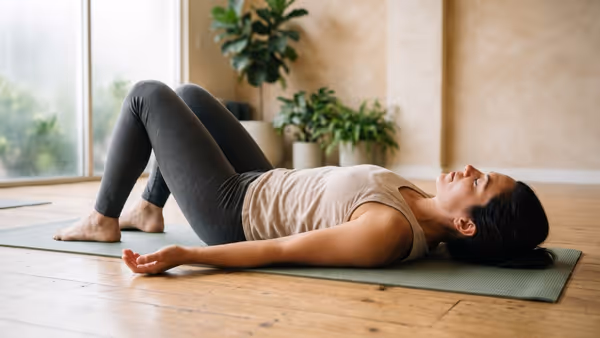 A woman lying on her back on a sage-green mat on a wooden floor, knees bent with both feet flat, one arm resting out to the side with the palm up, eyes closed and face relaxed, seen from floor level