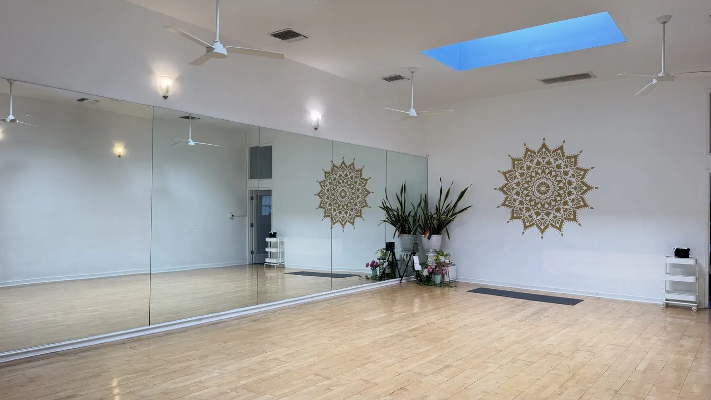 The practice room seen wide, a pale wood floor running to a full mirrored wall, two gold mandala discs on the facing wall, ceiling fans and two blue skylights overhead