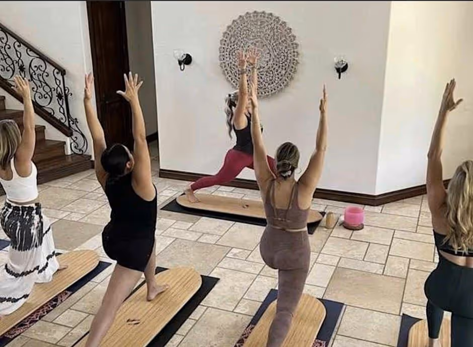 Photograph of a surf yoga class indoors: five women in a high lunge with arms raised, each standing on a wooden surfboard-shaped balance board laid on a mat, on a tiled floor beside a staircase