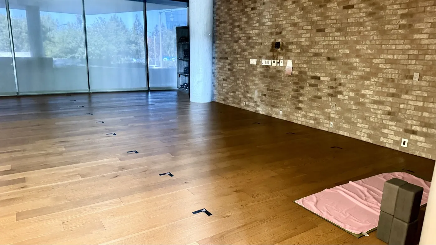A second practice room with a wood floor, a brick wall along one side and windows along the other, one pink mat and two blocks left out on the floor.