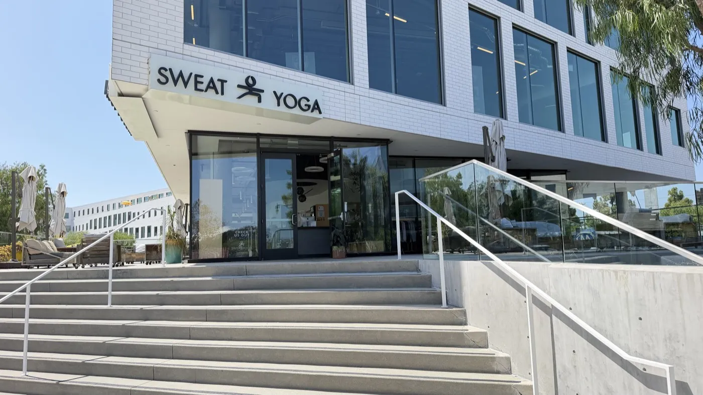 The entrance to Sweat Yoga Playa Vista: a SWEAT YOGA sign above glass doors at the top of a wide concrete staircase, on the ground floor of a white office building.
