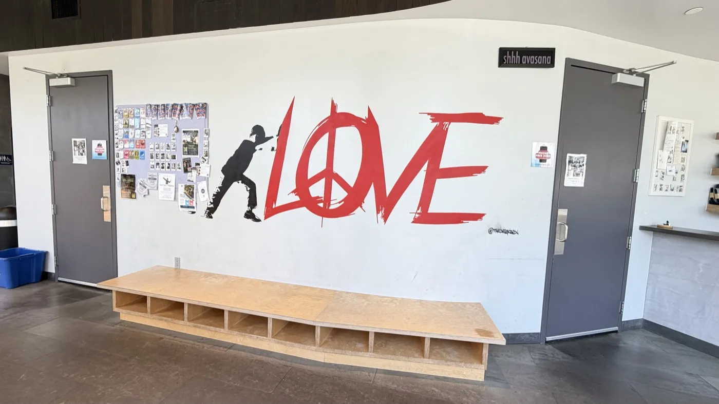 A red LOVE mural on a white wall between two studio doors, with a long wooden bench and cubby shelf beneath it and a small shhh savasana sign above one door.