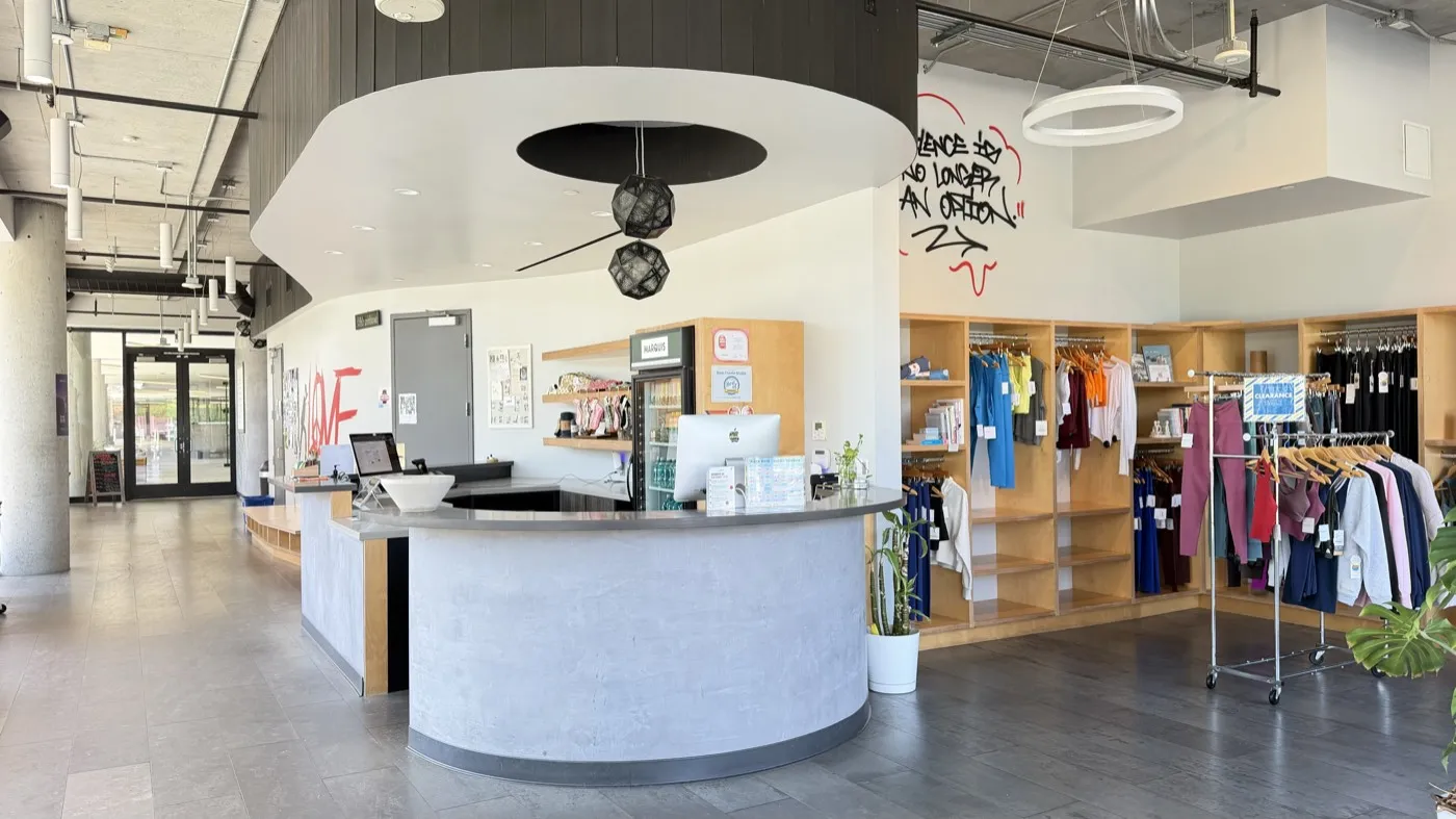 Reception at Sweat Yoga Playa Vista: a curved concrete desk under a round white canopy, with clothing rails and shelving to one side and the entrance doors behind.