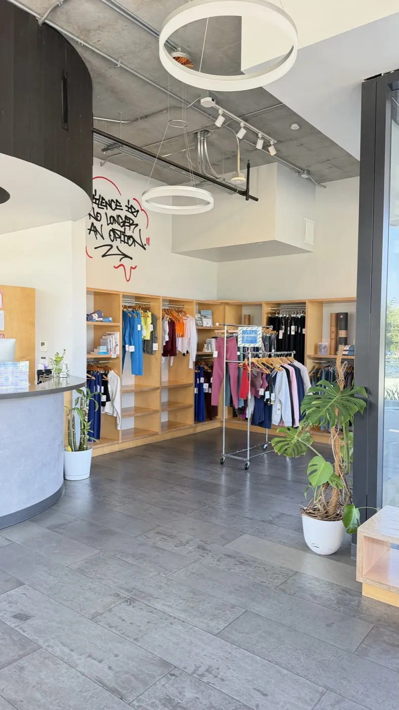 The retail corner at Sweat Yoga Playa Vista: wooden shelving of folded clothing, rails of leggings and tops, a large monstera plant and the reception desk to the left.