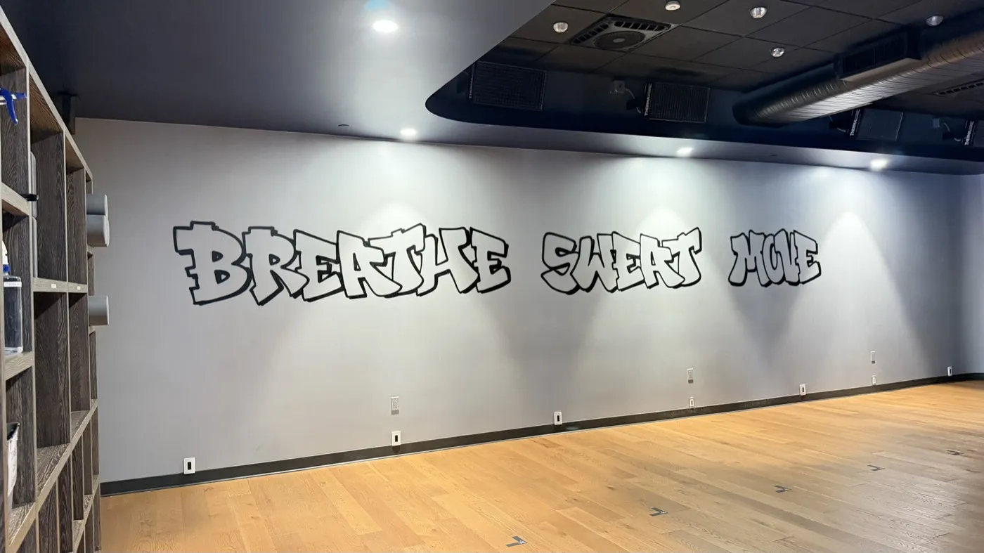 A studio wall painted with the words BREATHE SWEAT MOVE in black graffiti lettering, lit by downlights, with exposed ducting overhead.