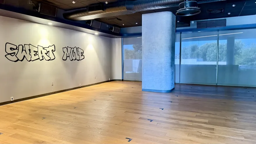 A practice room at Sweat Yoga Playa Vista: pale wood floor, a structural pillar lit blue, frosted windows down one side and graffiti lettering across the left wall.