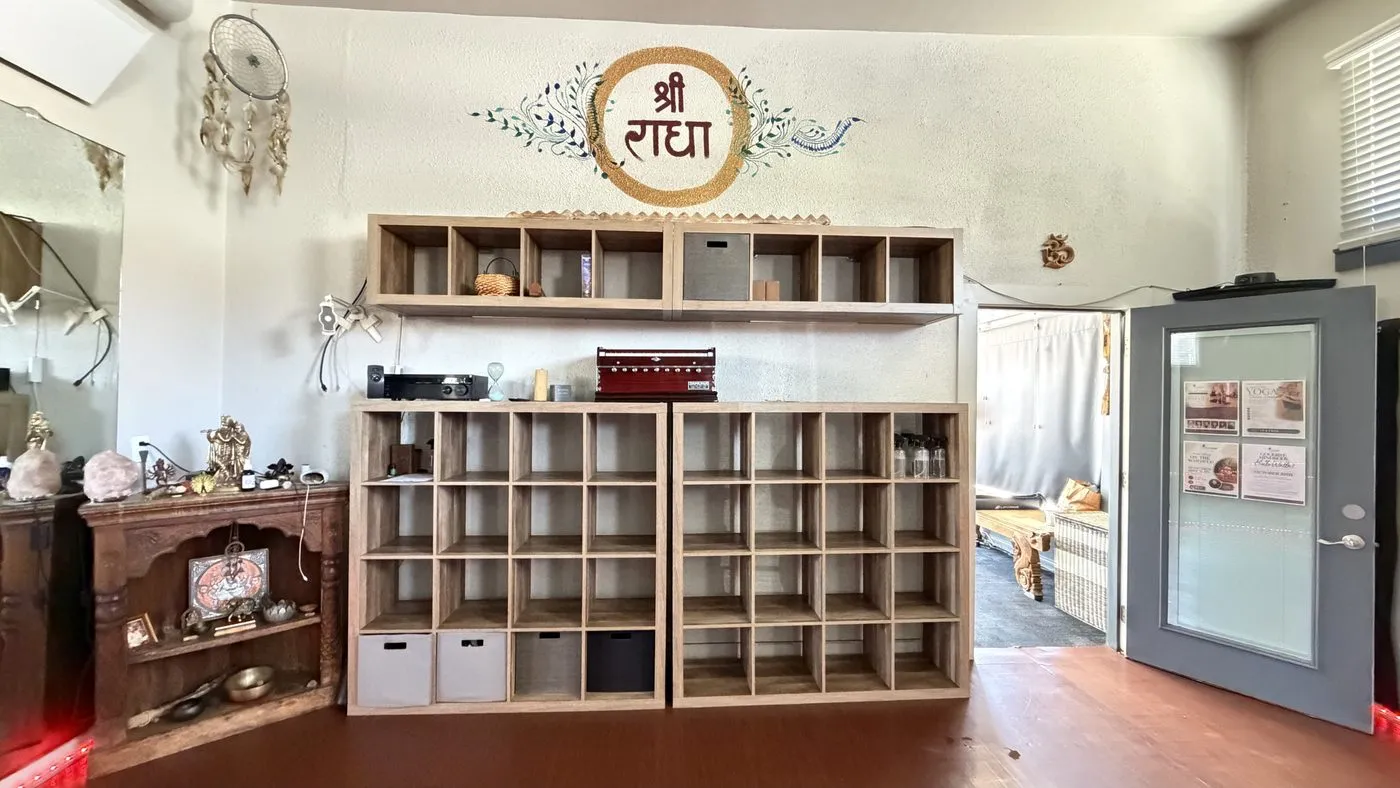 The cubby wall, two tiers of open wooden pigeonholes for shoes and bags under a painted roundel reading Shri Radha, with a dreamcatcher and a small shrine alongside