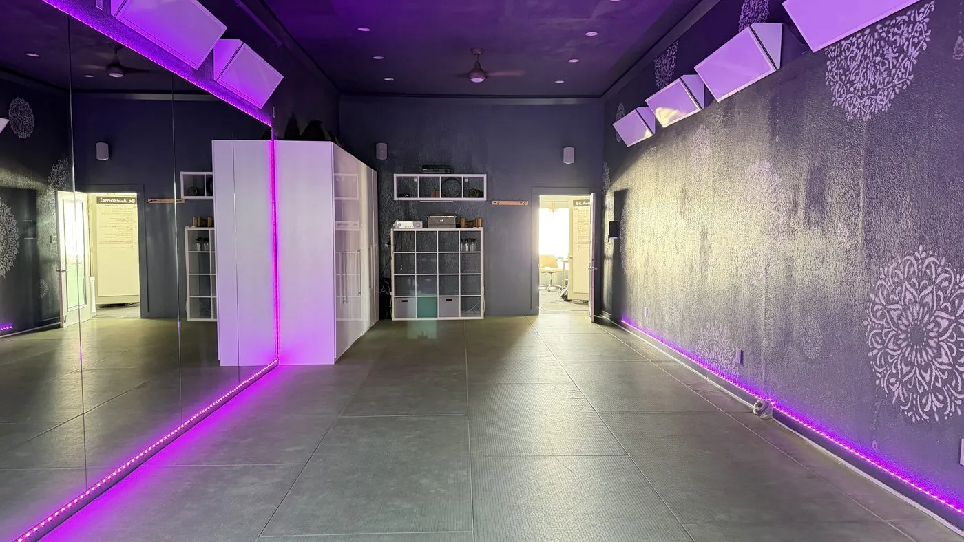 The heated room, walls and ceiling in deep purple with magenta light strips along the floor and ceiling, large mandala stencils on the wall and prop shelving at the far end