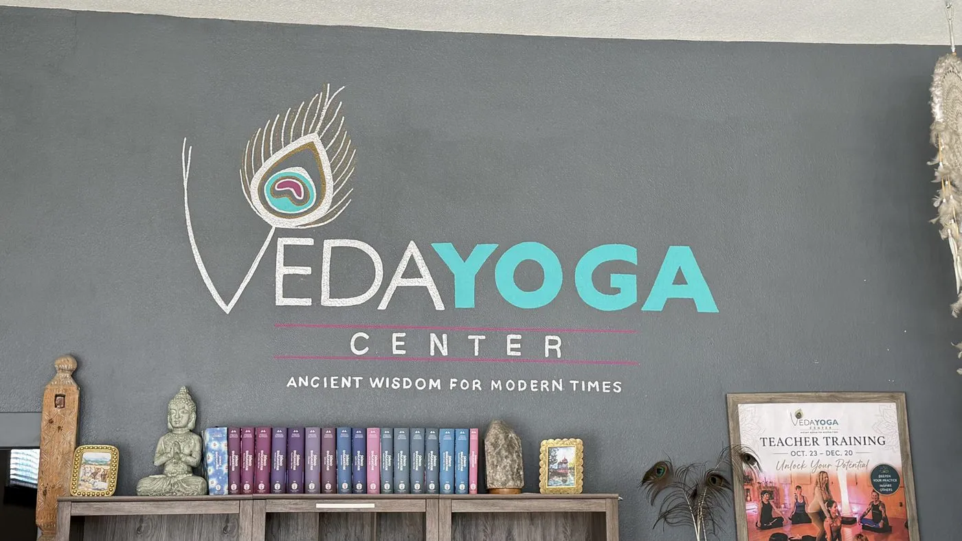 The Veda Yoga Center peacock-feather logo painted on a grey wall above the line ancient wisdom for modern times, with a low console of books and a seated Buddha beneath