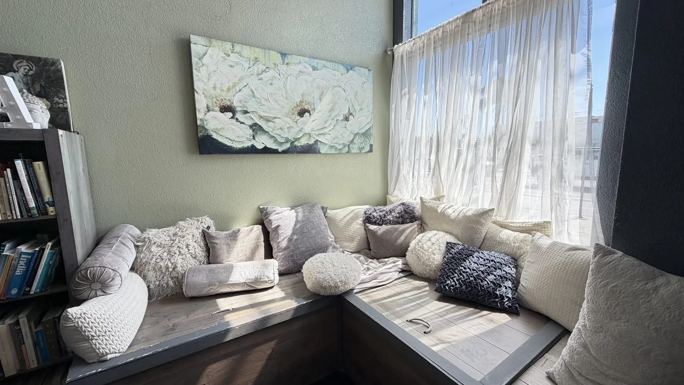 The lounge, an L-shaped built-in window seat heaped with cream and grey cushions beside a bookshelf, sheer curtains and a white-flower canvas on the wall