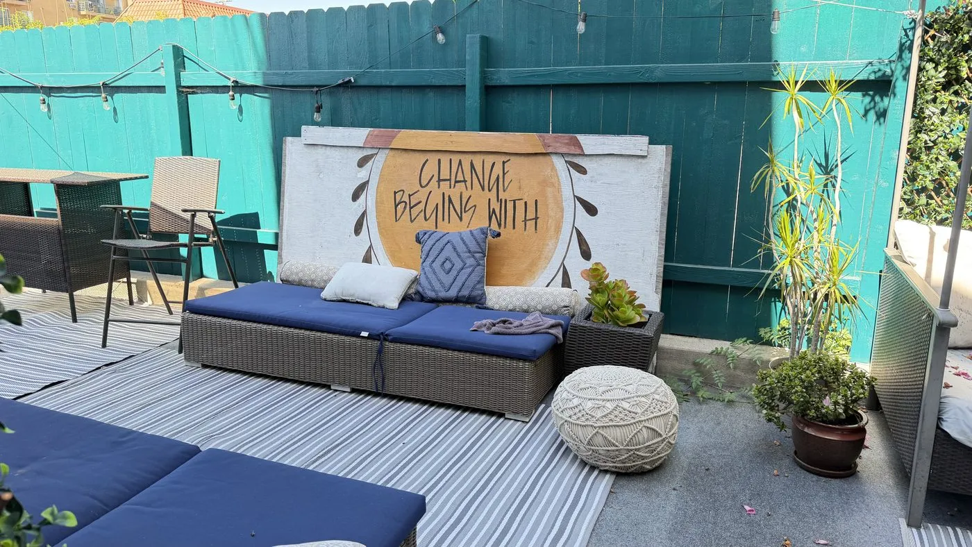 The outdoor patio, a navy-cushioned daybed against a teal fence hung with string lights, a painted sign reading change begins with, and potted bamboo alongside