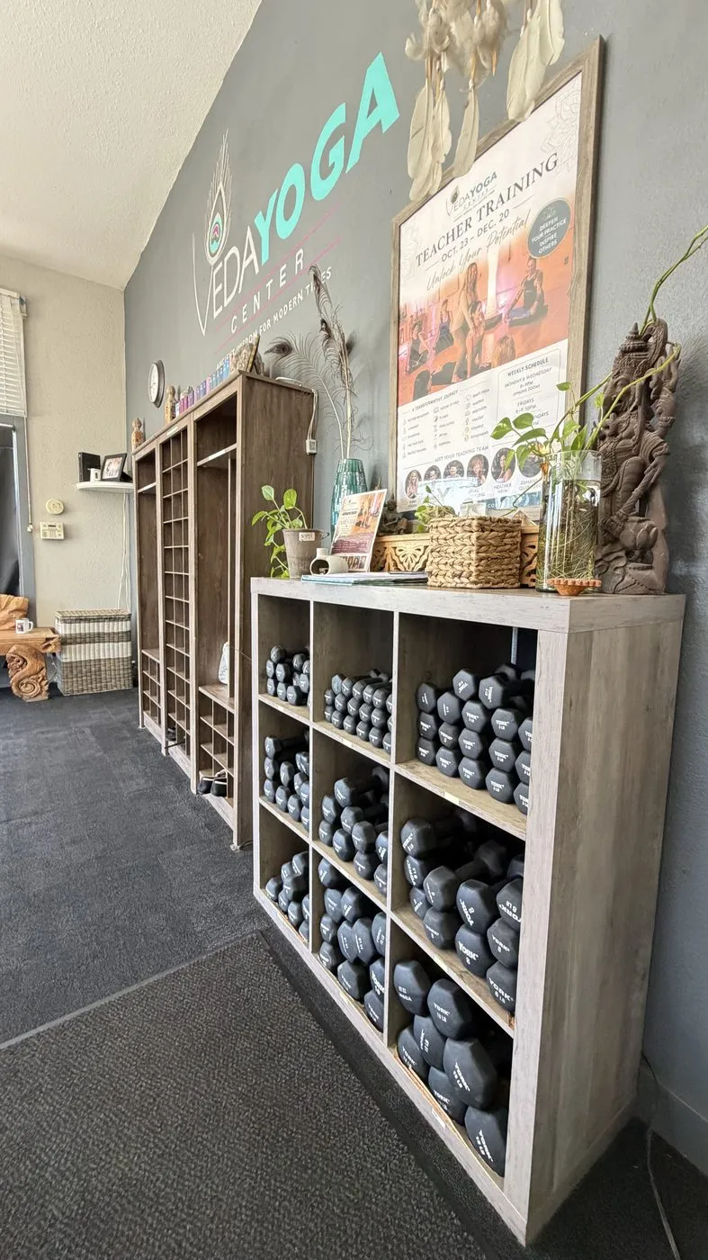 Prop shelving stacked with grey blocks and rolled bolsters, beside a wall carrying the Veda Yoga logo and a teacher-training poster