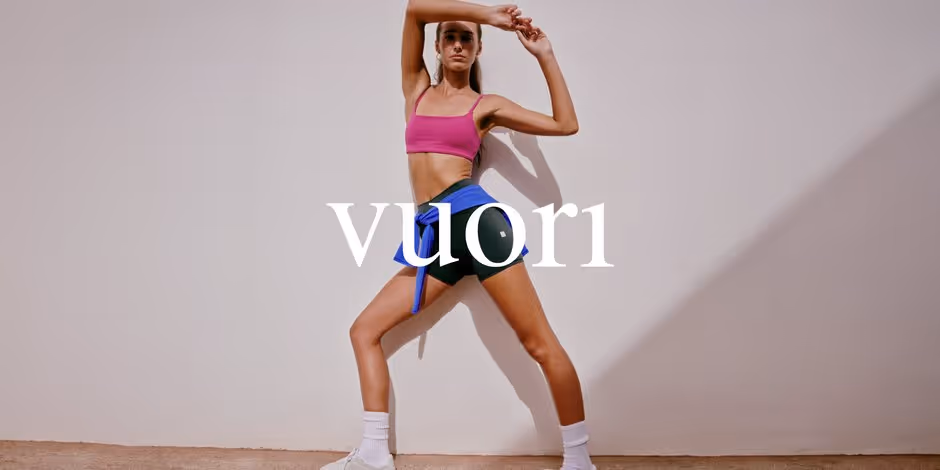Promotional banner for the event: a woman in a magenta bralette and dark green bike shorts, a blue top tied at her waist, stands in a wide stance with both arms raised against a sunlit off-white wall, with the white vuori wordmark across the middle