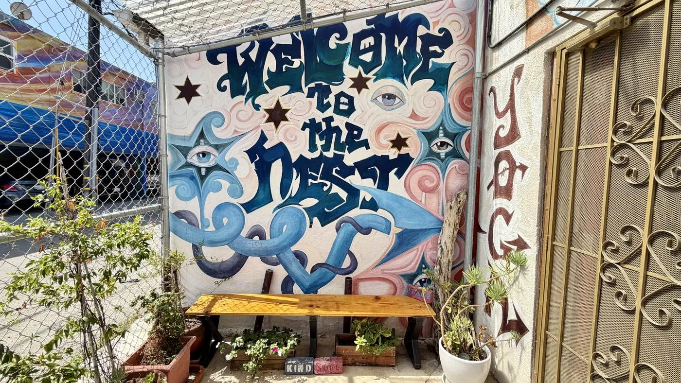 The entrance: a mural reading WELCOME TO THE NEST in blue lettering among painted eyes and waves, with a wooden bench and potted plants beneath it