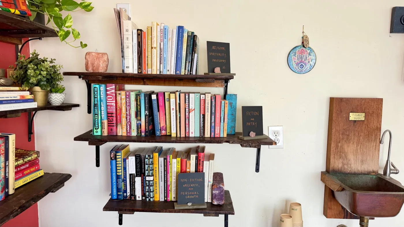 The lending library from the side, beside a carved wooden basin plaqued Filtered Water with a stack of paper cups under the tap