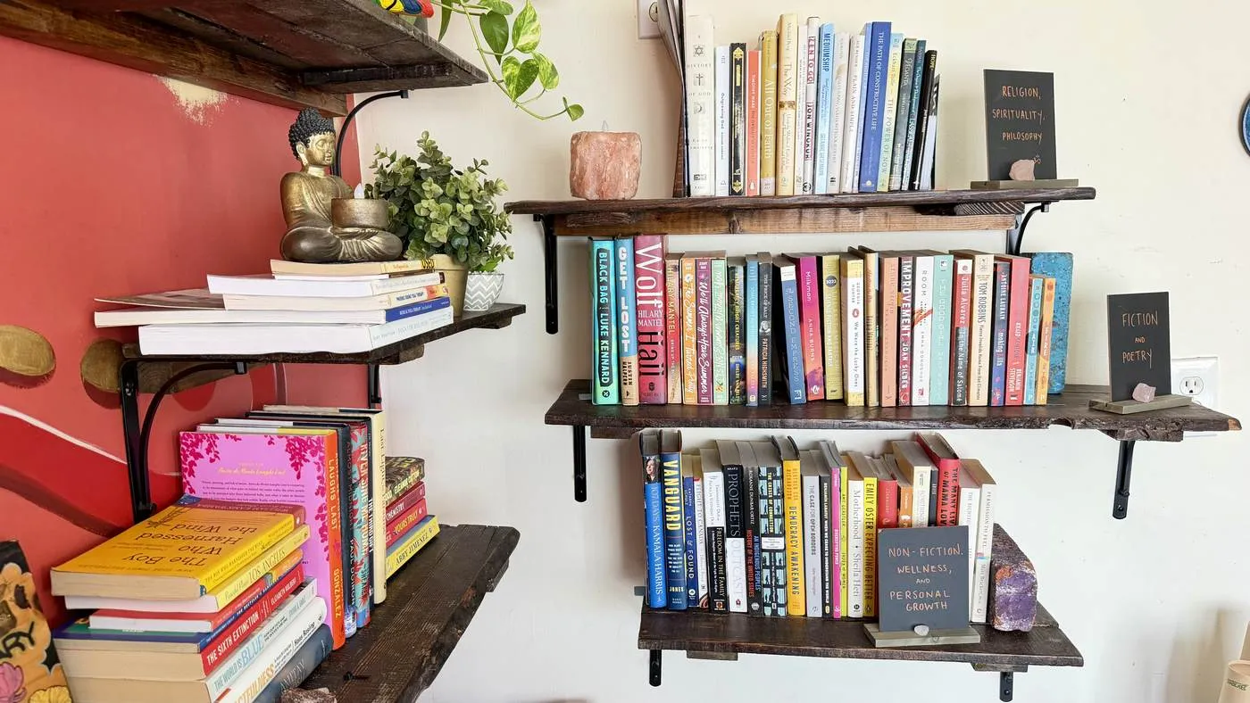 A wall-mounted lending library on live-edge shelves, its sections hand-lettered on slate cards — religion and philosophy, fiction and poetry, non-fiction and wellness — with a brass Buddha and crystals among the books