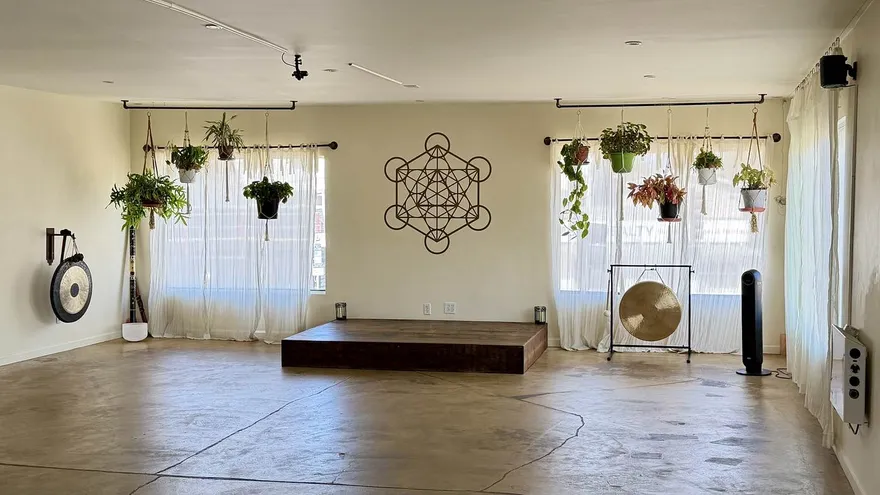 The main practice room: a polished concrete floor, sheer curtains over tall windows, hanging plants along both walls, a Metatron's Cube decal and a low wooden platform with a brass gong beside it